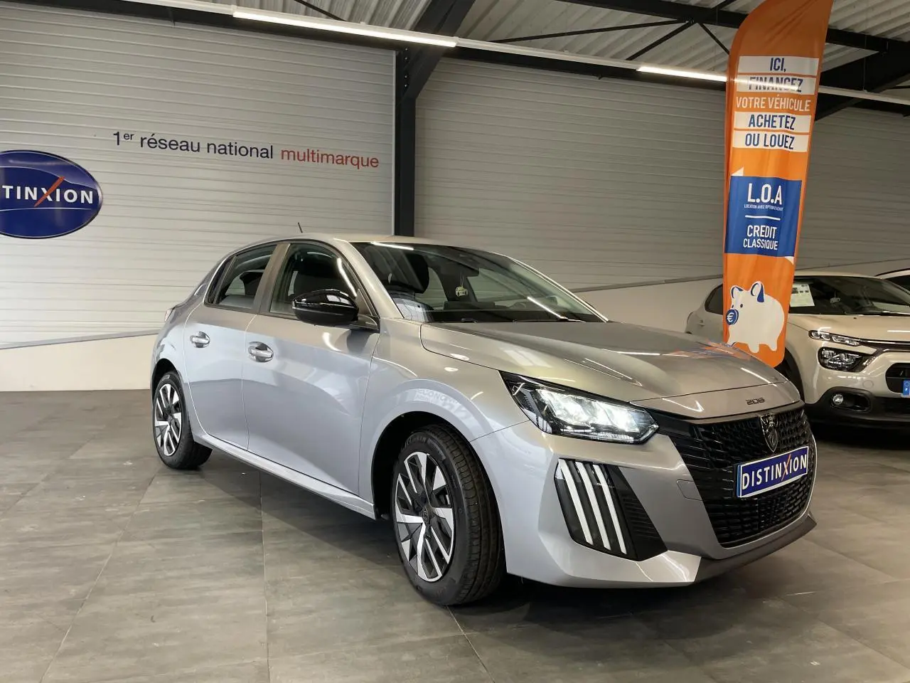 Peugeot 208 gris clair vue 3/4 avant droit en intérieur, avec calandre noire et feux LED distinctifs.