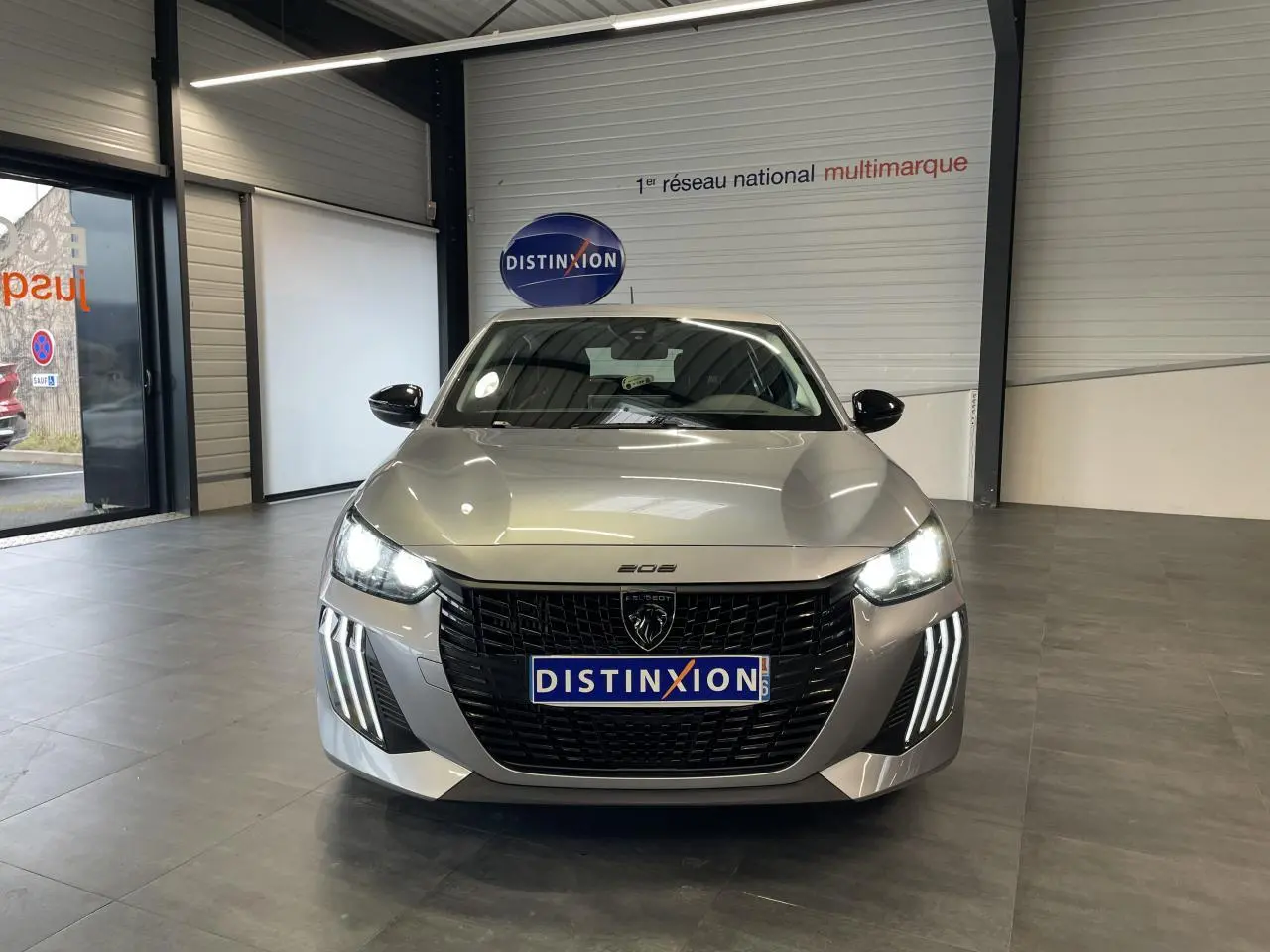 Vue frontale d'une Peugeot 208 gris clair avec calandre noire et feux LED à griffes allumés en intérieur showroom.