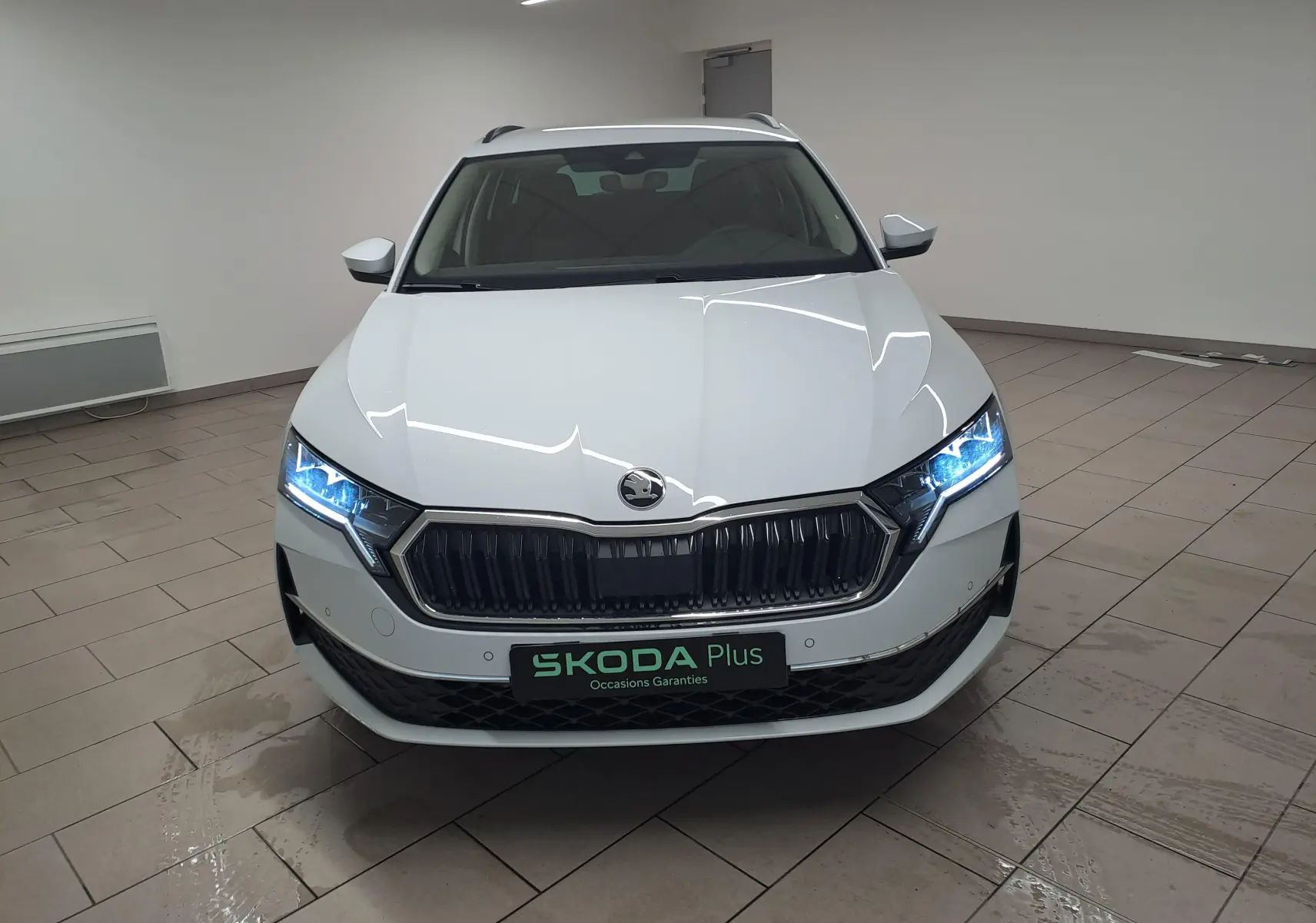 Vue frontale d'une Skoda Octavia Combi 2.0 TDI blanche Moon avec phares allumés dans un intérieur neutre.