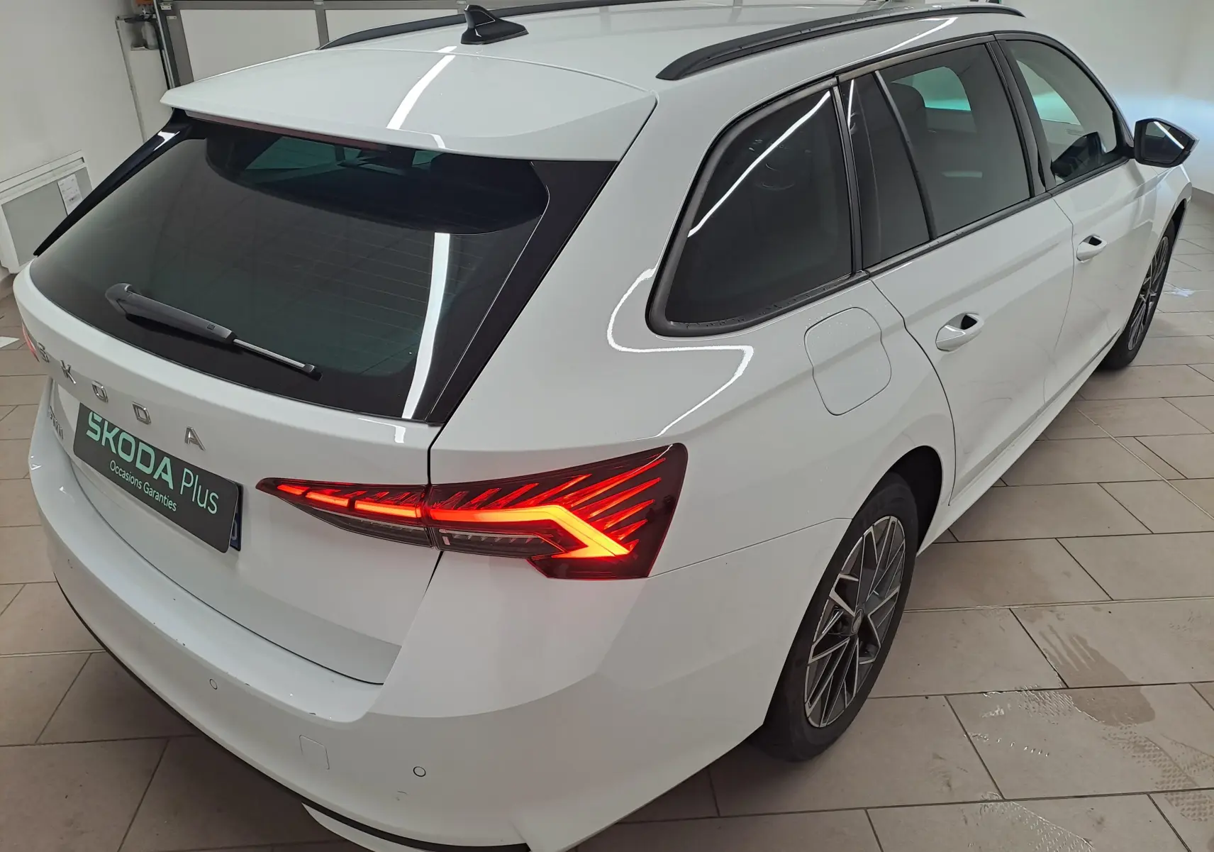 Vue 3/4 arrière droite d'une Skoda Octavia Combi blanche Moon avec feux arrière allumés et jantes alliage noires polies.