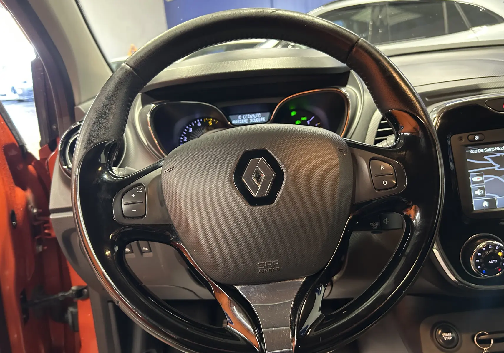 Volant noir brillant avec logo Renault au centre, tableau de bord et écran GPS visibles dans un Renault Captur orange 2014.