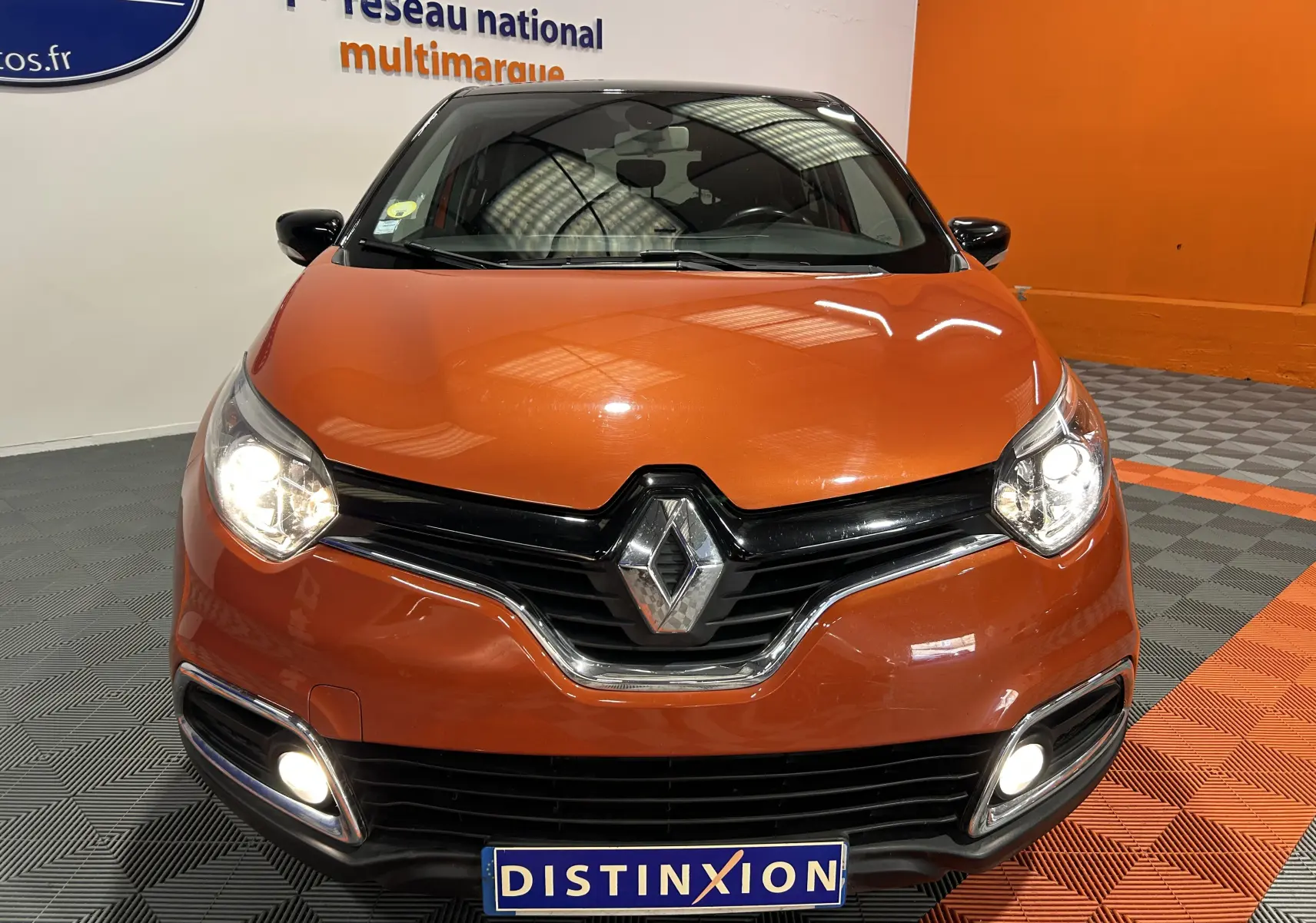 Vue frontale d'un Renault Captur orange 2014 avec phares et feux antibrouillard allumés en intérieur showroom