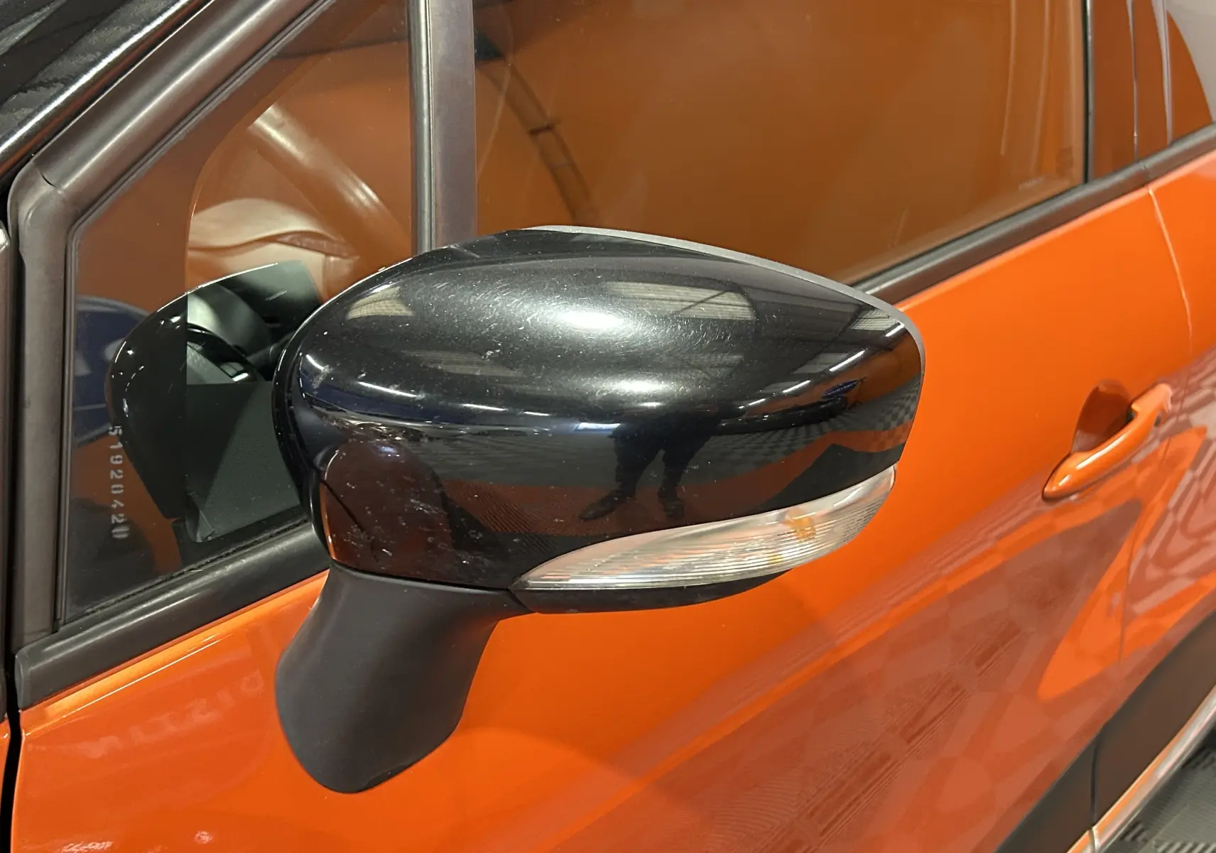 Rétroviseur noir en gros plan côté gauche d'un Renault Captur orange 2014 avec portière visible.