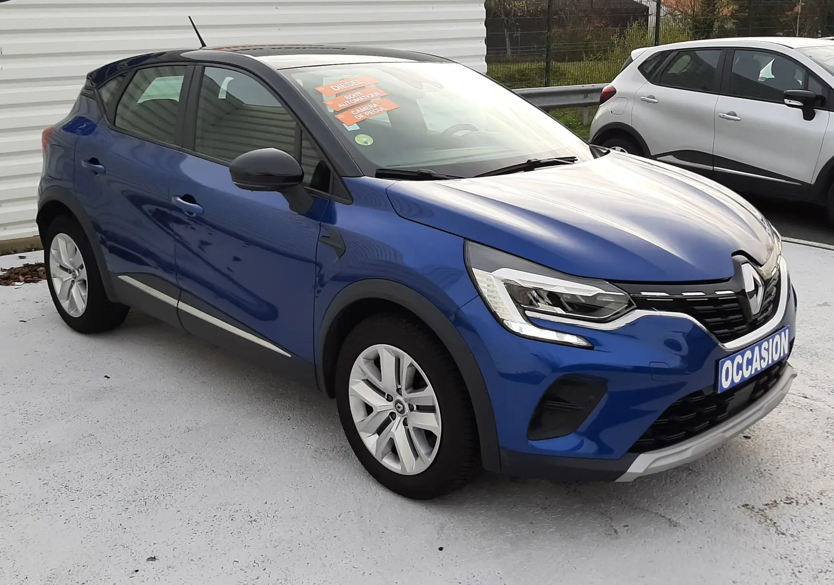 Renault Captur bleu Iron avec toit noir, vue 3/4 avant droit, stationné devant un panneau multimarque.