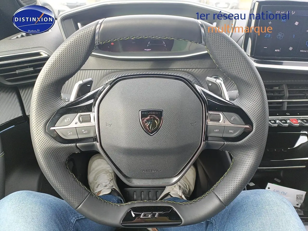 Vue rapprochée du volant cuir perforé avec logo Peugeot et badge GT, tableau de bord et écran tactile en arrière-plan.