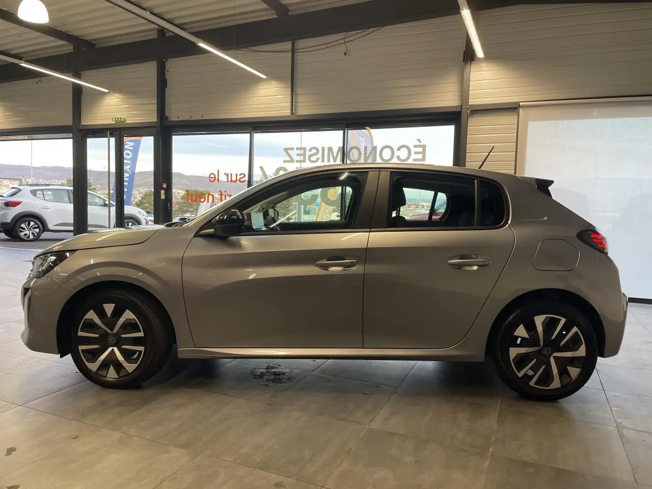 Profil droit d'une Peugeot 208 2024 gris clair avec jantes 16'' et feux arrière à LED en intérieur showroom.