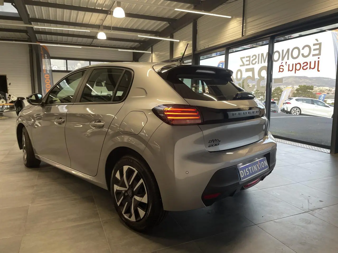 Vue 3/4 arrière droite d'une Peugeot 208 2024 gris clair avec feux arrière à LED et jantes stylisées en concession.
