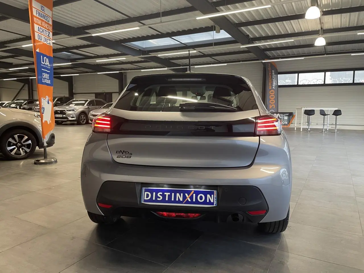 Vue arrière d'une Peugeot 208 gris clair 2024 avec feux arrière à LED et bandeau noir brillant en concession.
