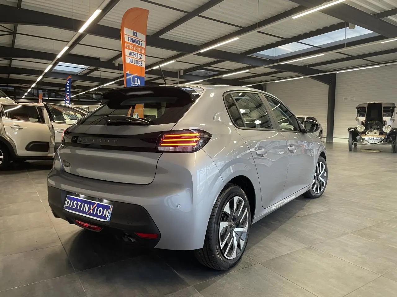 Vue 3/4 arrière droite d'une Peugeot 208 2024 gris clair métallisé avec feux arrière à LED et jantes stylisées.