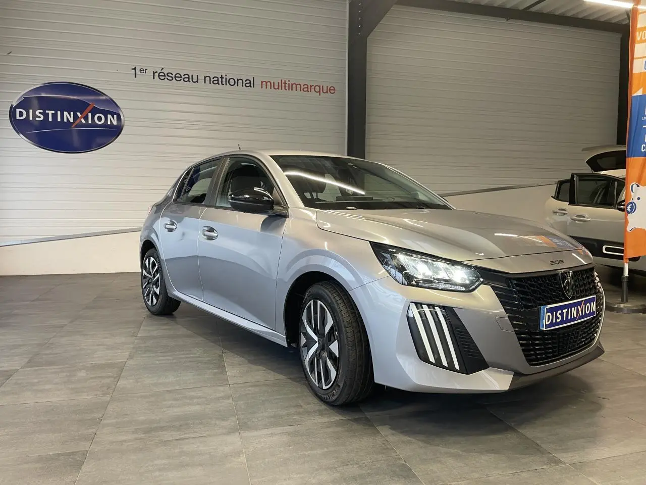 Peugeot 208 gris clair vue 3/4 avant droit en intérieur avec calandre noire et feux LED griffés.