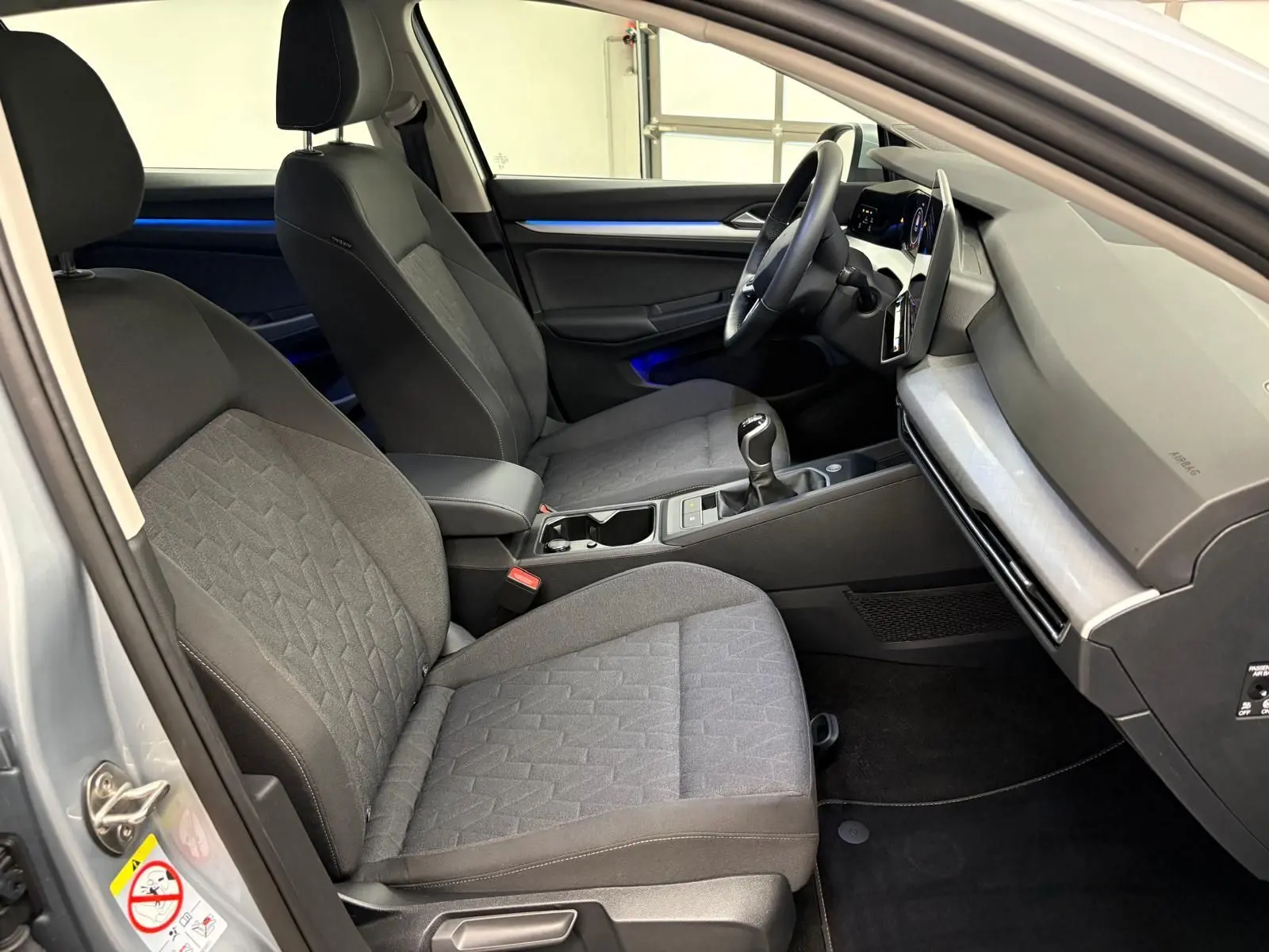 Vue intérieure côté conducteur de la Volkswagen Golf 2.0 TDI 2025 en reflet d'argent, avec sellerie tissu et éclairage d'ambiance bleu.