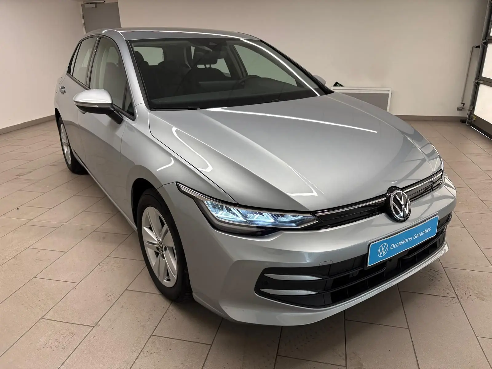 Volkswagen Golf 2.0 TDI 2025 en reflet d'argent, vue 3/4 avant droit avec phares LED allumés en intérieur.