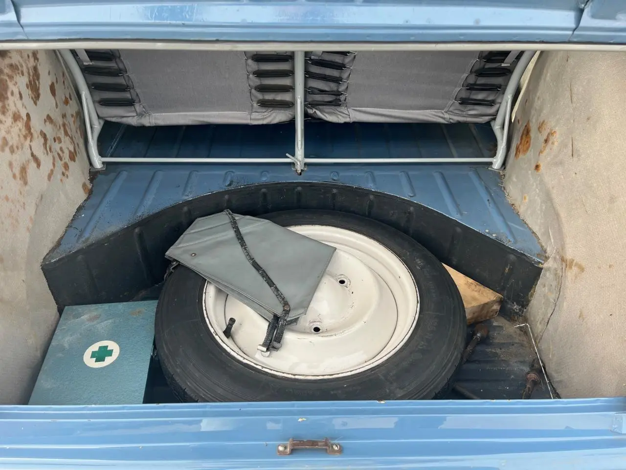 Coffre ouvert d'une Citroën 2 CV bleu clair de 1960, montrant la roue de secours et une trousse de secours.