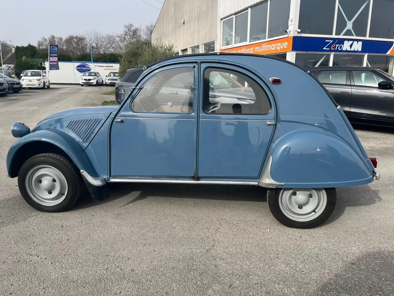 Profil droit d'une Citroën 2 CV bleu clair de 1960, avec ses roues étroites et sa carrosserie arrondie caractéristique.