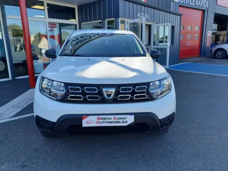 Vue avant d'un Dacia Duster blanc Glacier 2021 avec calandre noire et logo chromé, stationné devant un garage.