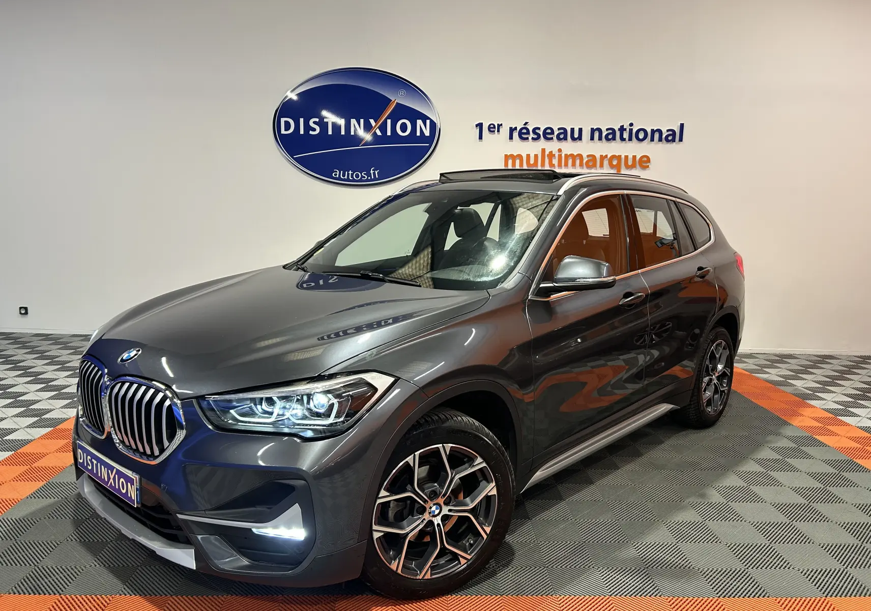BMW X1 gris en vue 3/4 avant droit, avec toit ouvrant et jantes alliage distinctives, dans un showroom lumineux.