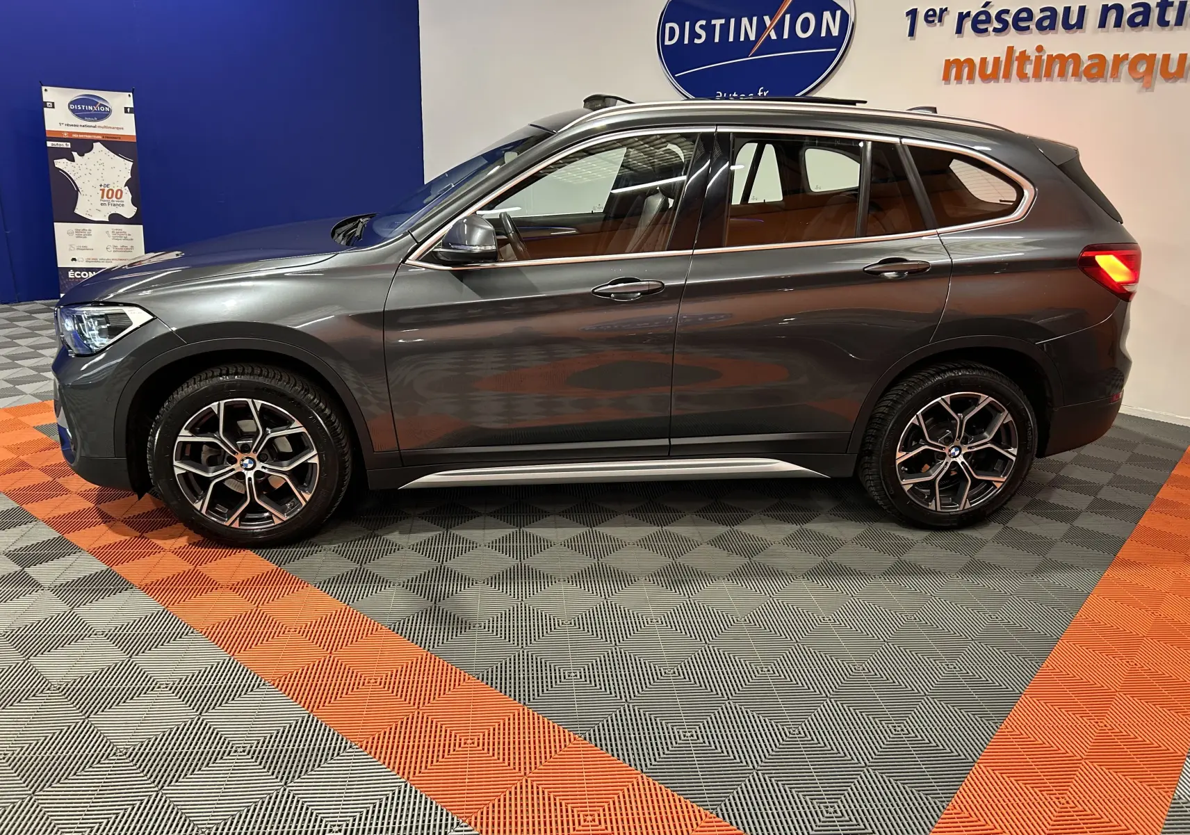 BMW X1 gris en vue profil côté gauche, avec jantes alliage et feux arrière allumés dans un showroom.