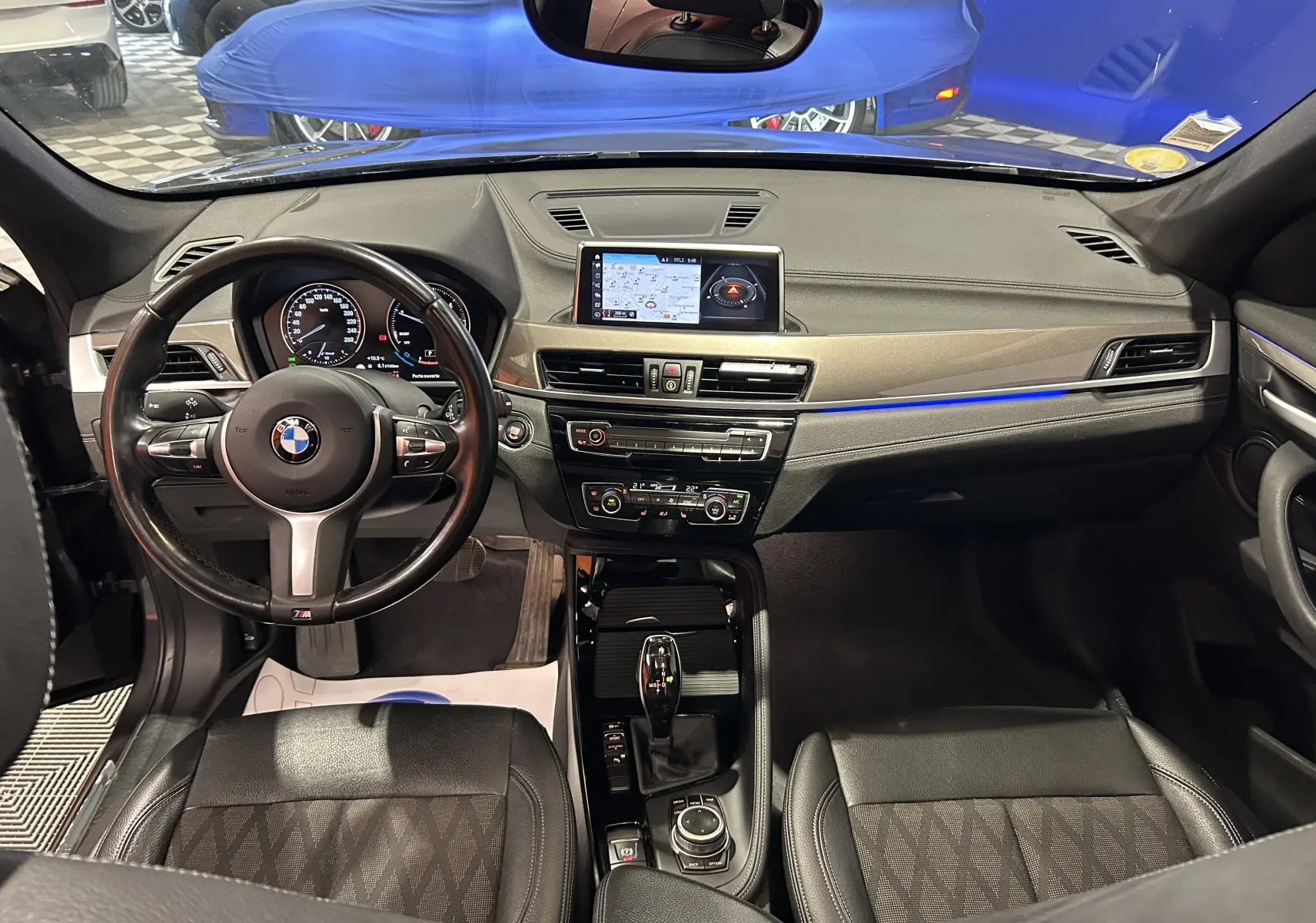 Vue intérieure avant du BMW X1 gris, mettant en valeur le tableau de bord avec écran central et éclairage d'ambiance bleu.