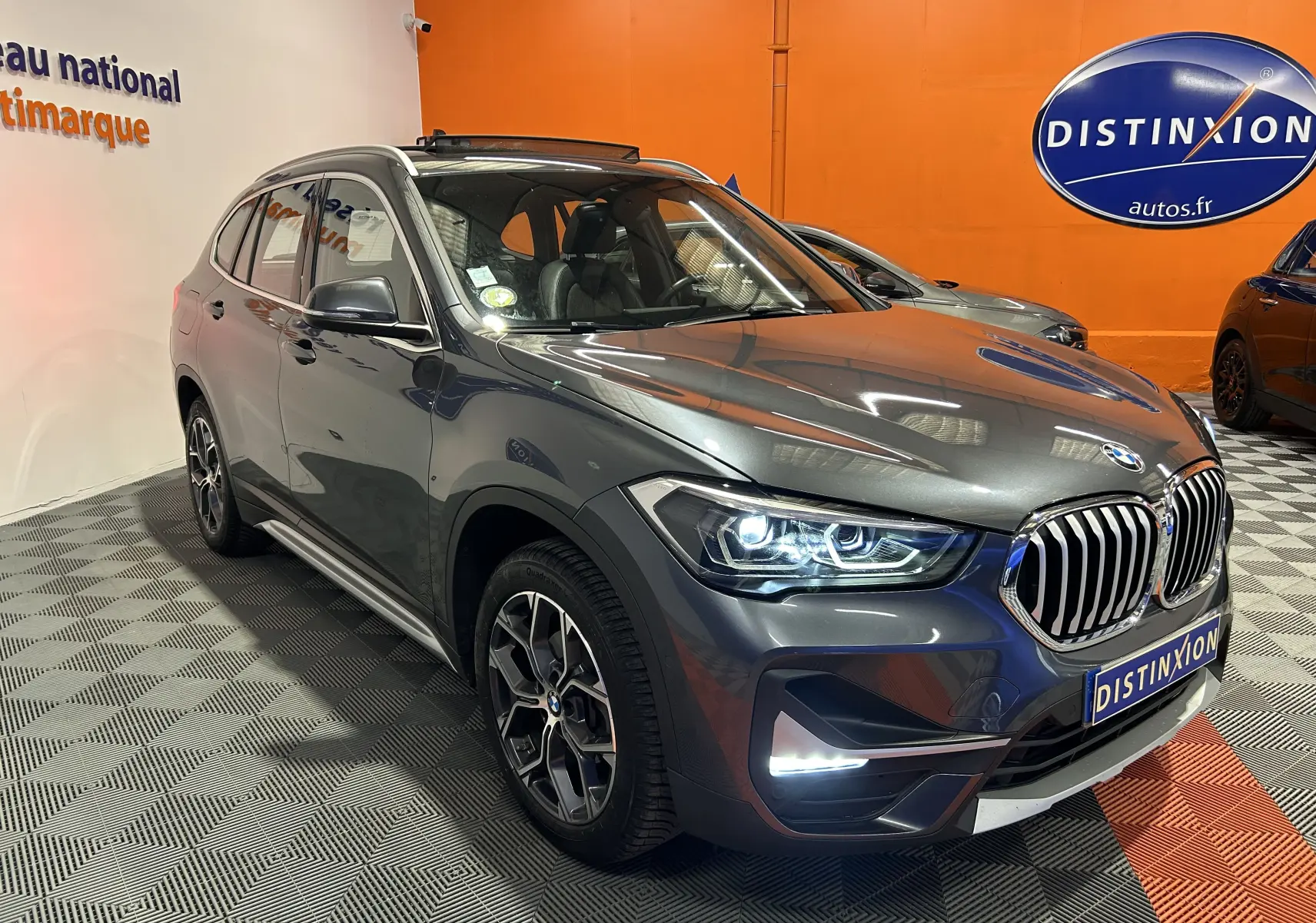 BMW X1 gris en 3/4 avant droit avec phares allumés et toit ouvrant visible en intérieur showroom.