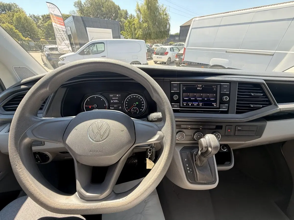 Vue intérieure centrée sur le volant et le tableau de bord du Volkswagen Transporter gris clair, avec écran tactile et levier de vitesses automatique.