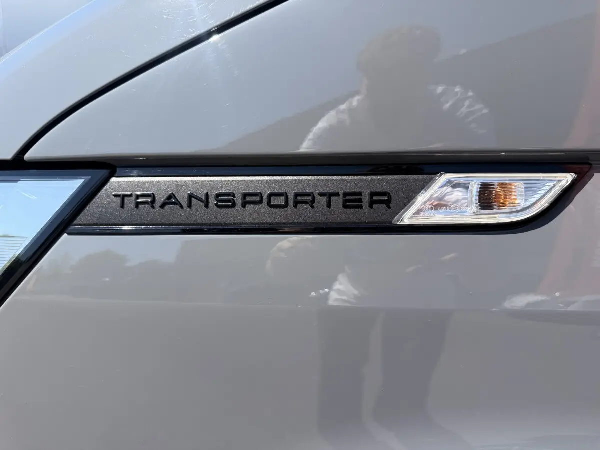 Détail du logo "Transporter" noir sur la porte gris clair d'un Volkswagen Transporter 2023, avec répétition lumineuse intégrée.