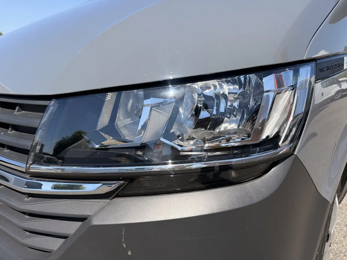 Gros plan sur le phare avant droit du Volkswagen Transporter gris clair, avec calandre et logo partiellement visibles.