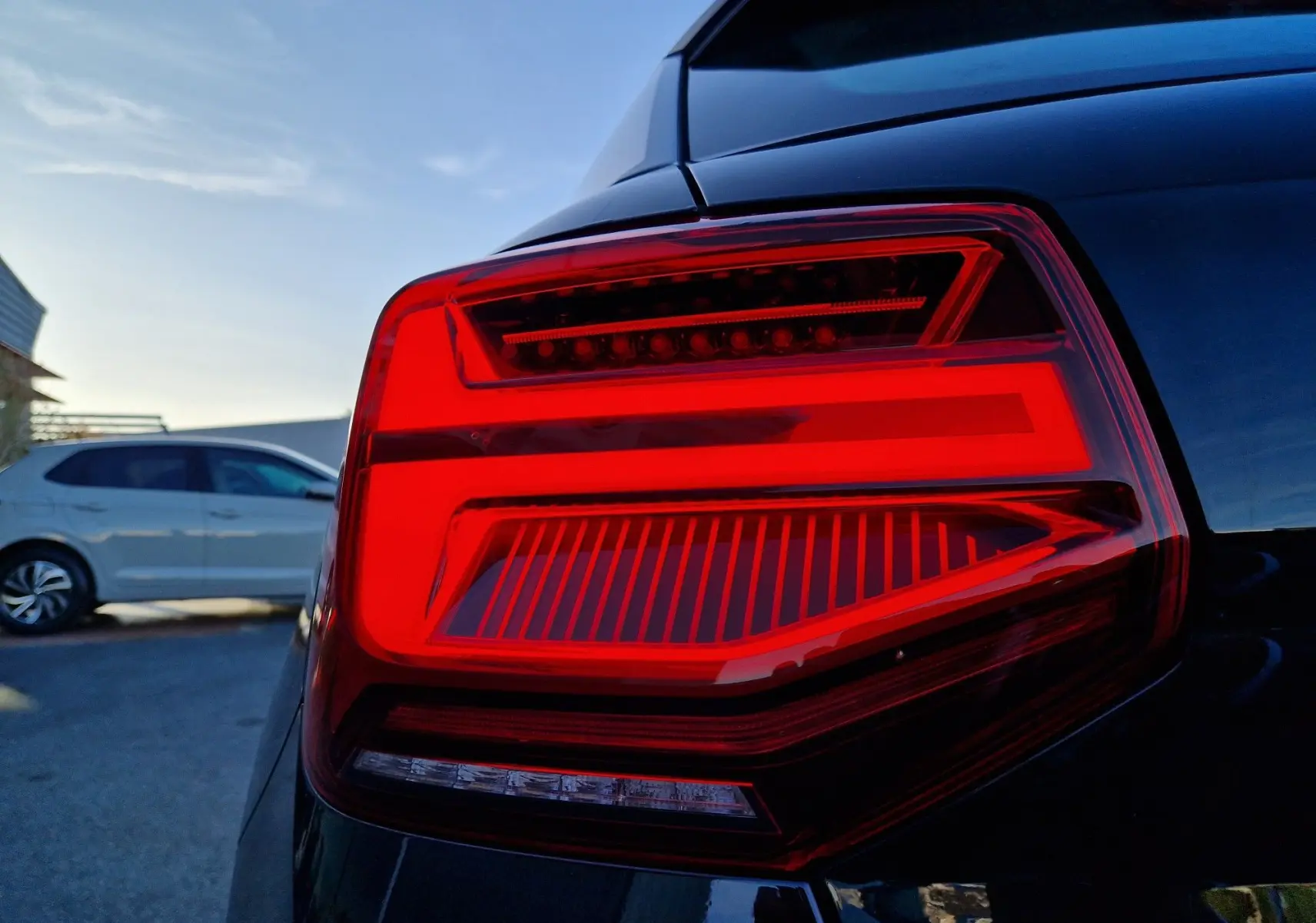 Gros plan sur le feu arrière droit rouge vif d’un Audi Q2 noir, avec un autre véhicule blanc en arrière-plan.