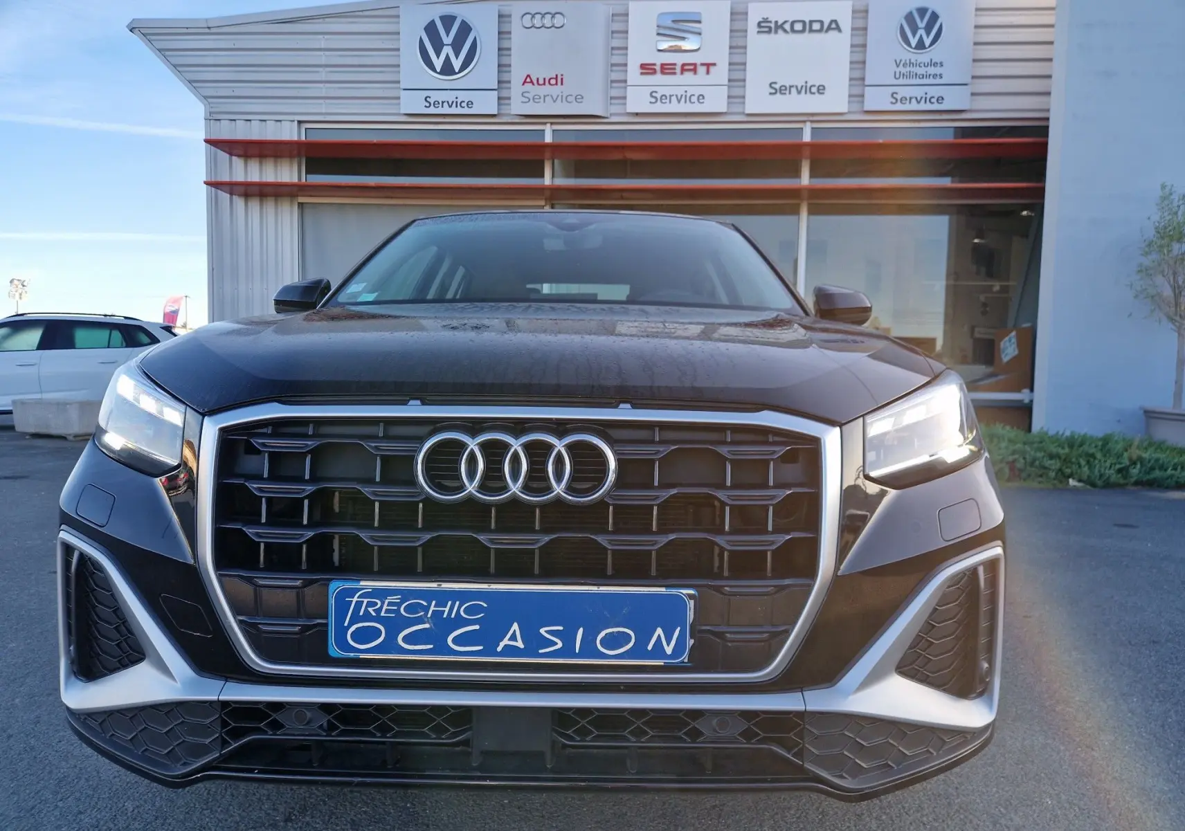 Vue frontale d’un Audi Q2 noir 2021 avec calandre large et phares LED allumés, devant un garage multimarques.