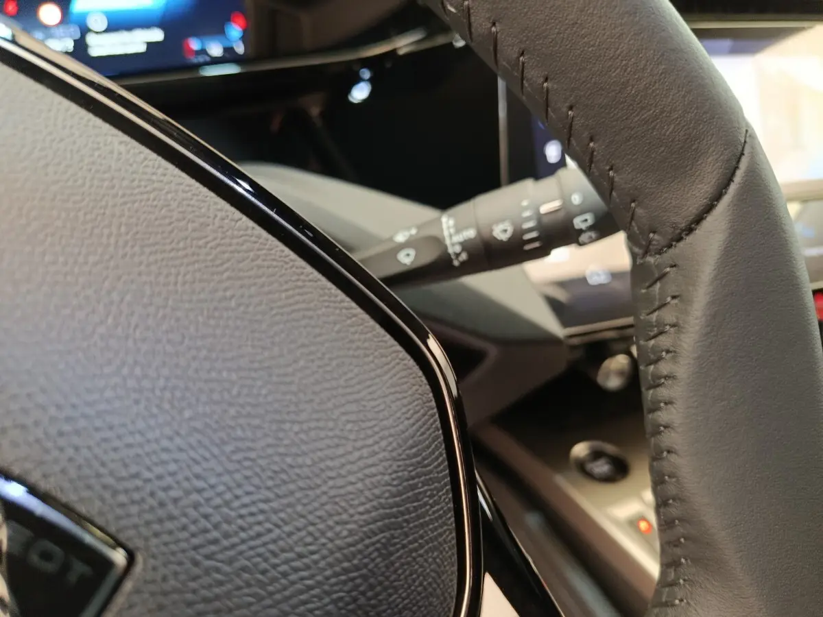 Gros plan sur le volant cuir noir multifonctions de la Peugeot 308 2026, avec tableau de bord et commandes visibles en arrière-plan.
