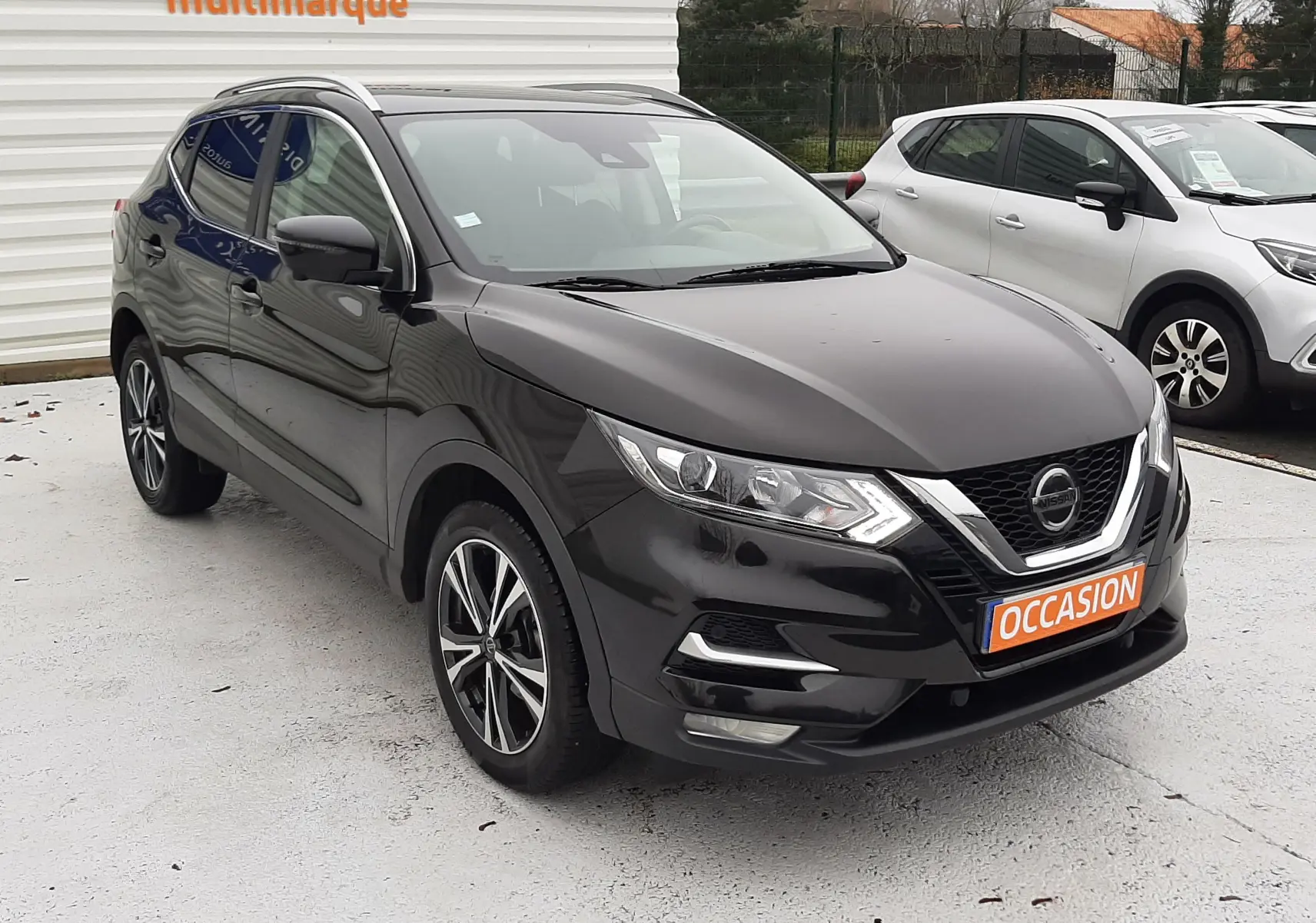 Nissan Qashqai noir en 3/4 avant droit, avec jantes alliage et plaque orange "OCCASION" visible sur un parking.