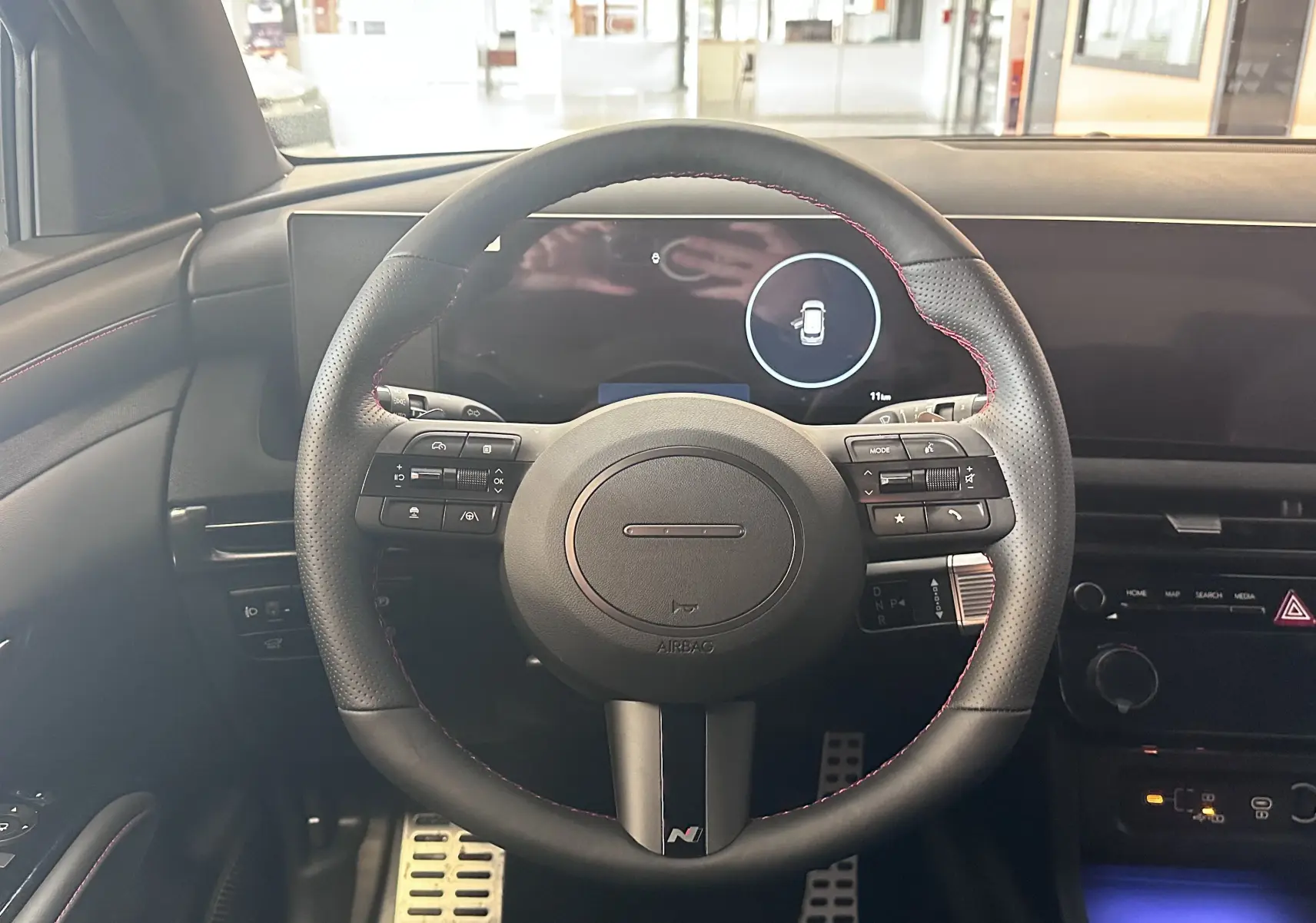 Vue intérieure centrée sur le volant noir avec surpiqûres rouges du Hyundai Tucson 2025, tableau de bord digital en arrière-plan.