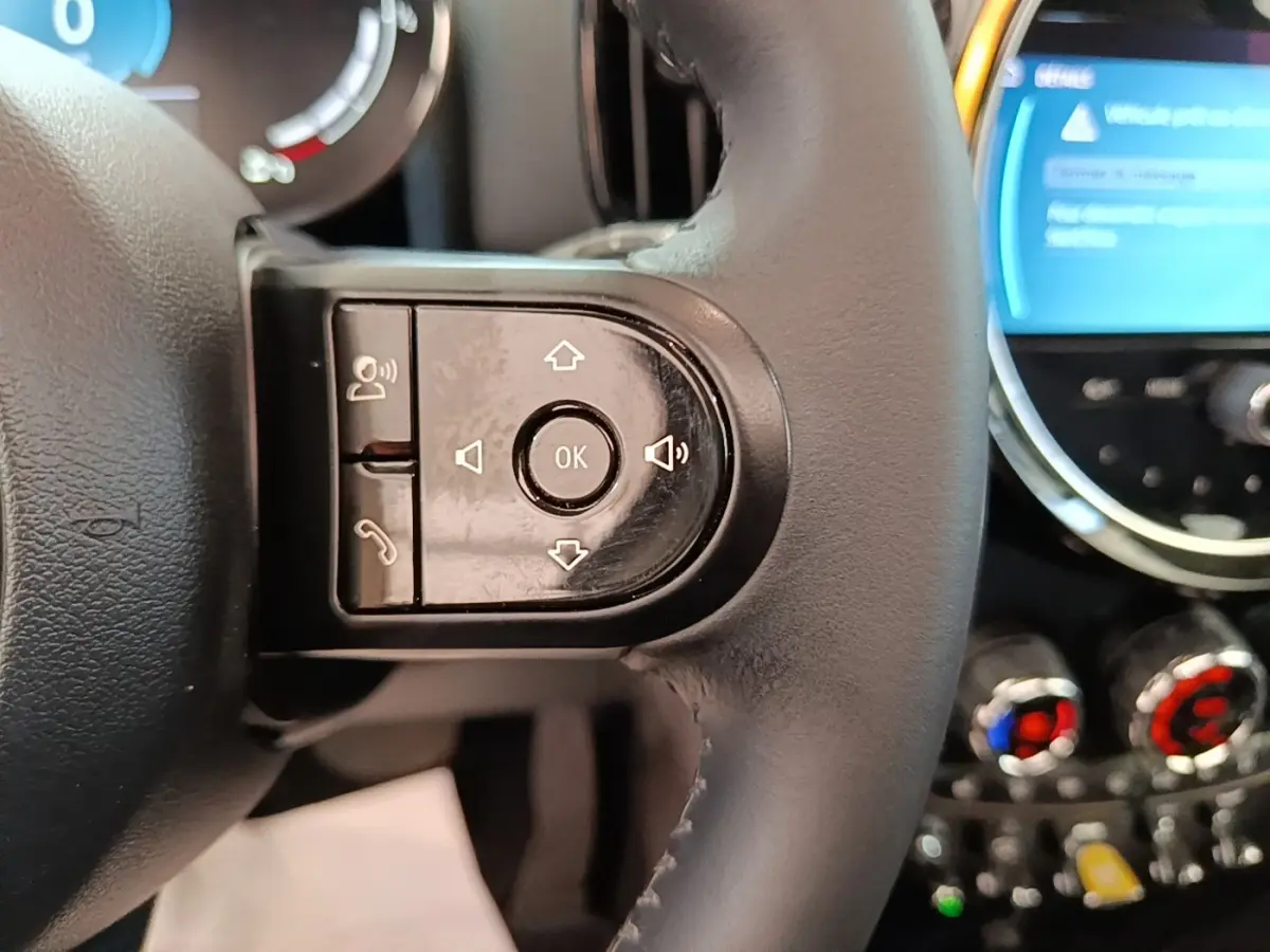 Gros plan sur le volant cuir noir multifonctions du MINI Countryman hybride, avec commandes vocales et écran tactile en arrière-plan.