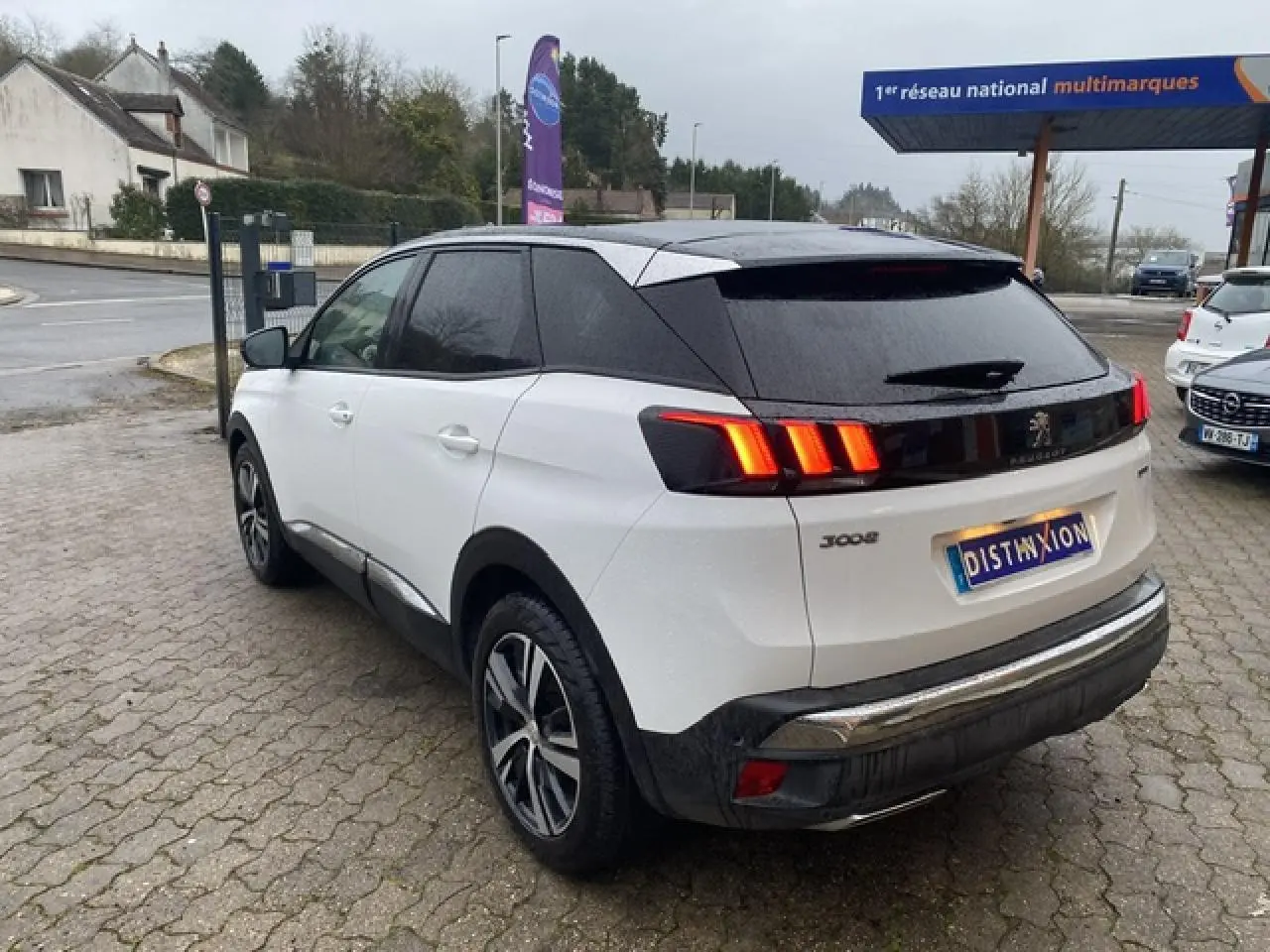 Vue 3/4 arrière droite d'un Peugeot 3008 blanc avec toit noir et feux arrière LED allumés sur parking.