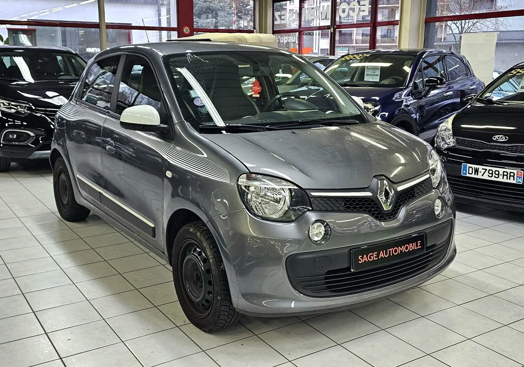 Renault Twingo III gris lunaire vue 3/4 avant droit en intérieur, avec rétroviseurs blancs et jantes acier noires.