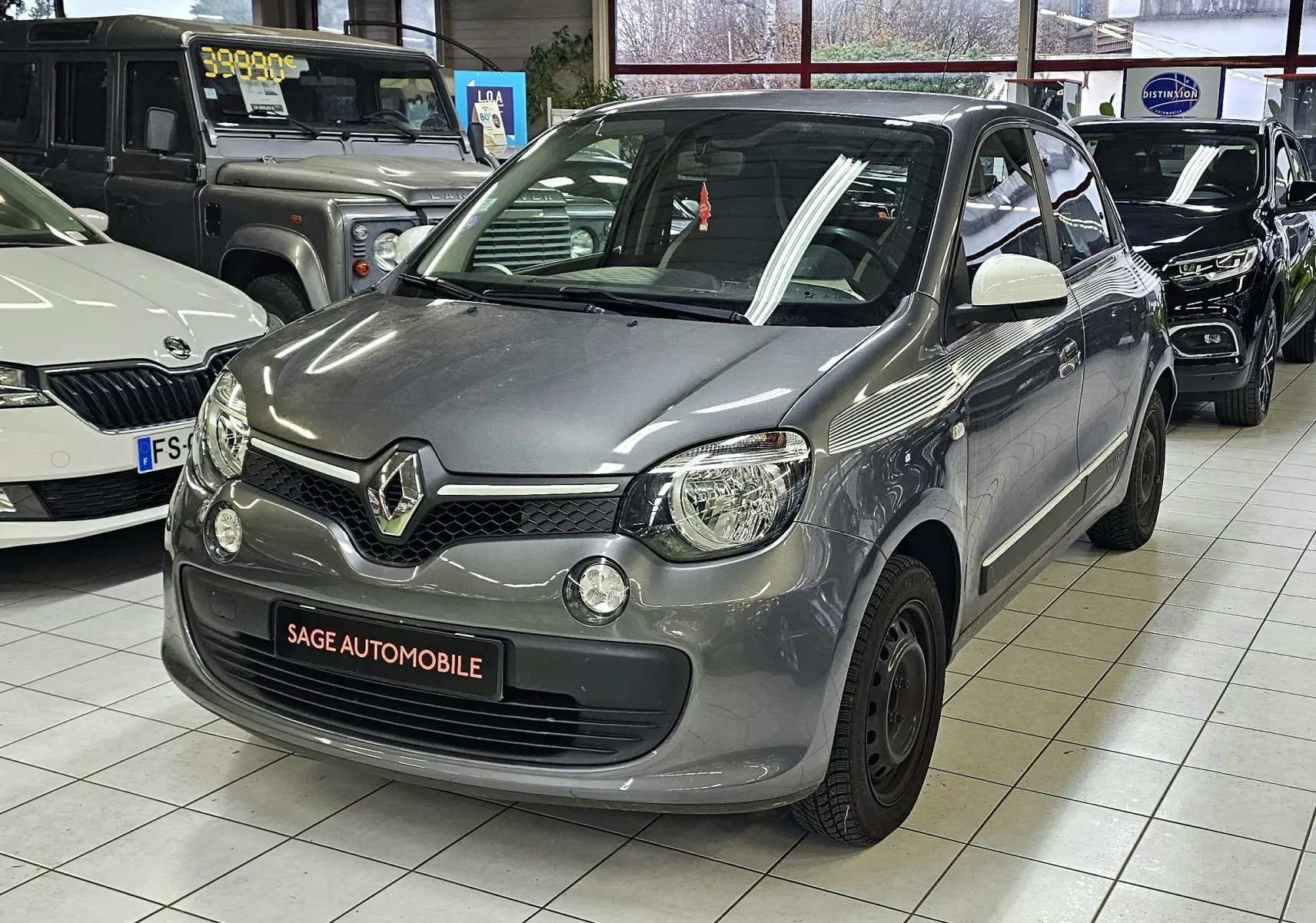 Renault Twingo III gris lunaire vue 3/4 avant droit en intérieur avec rétroviseurs blancs et jantes noires.