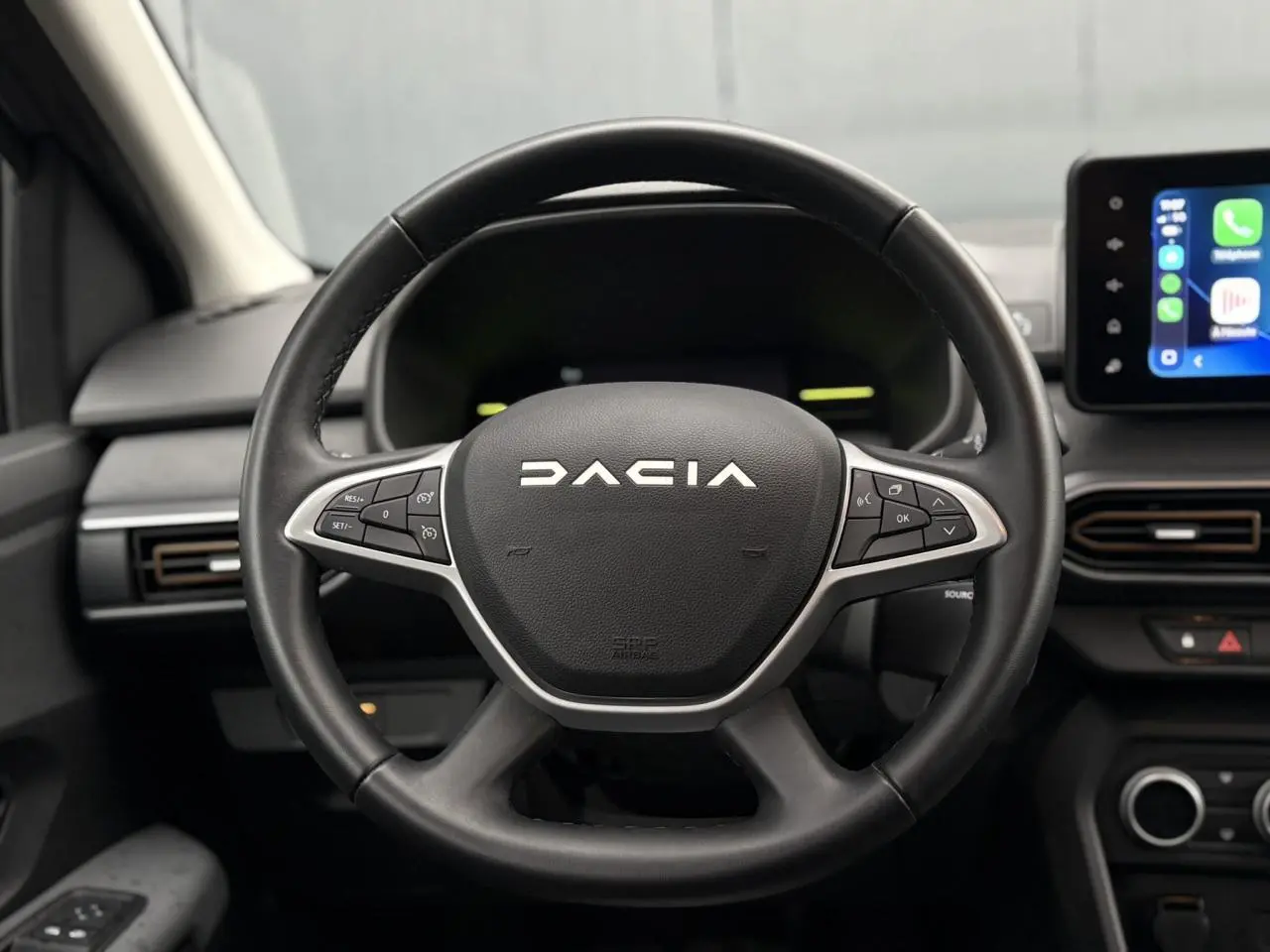 Vue rapprochée du volant noir DACIA avec commandes intégrées, tableau de bord et écran tactile à droite visibles.