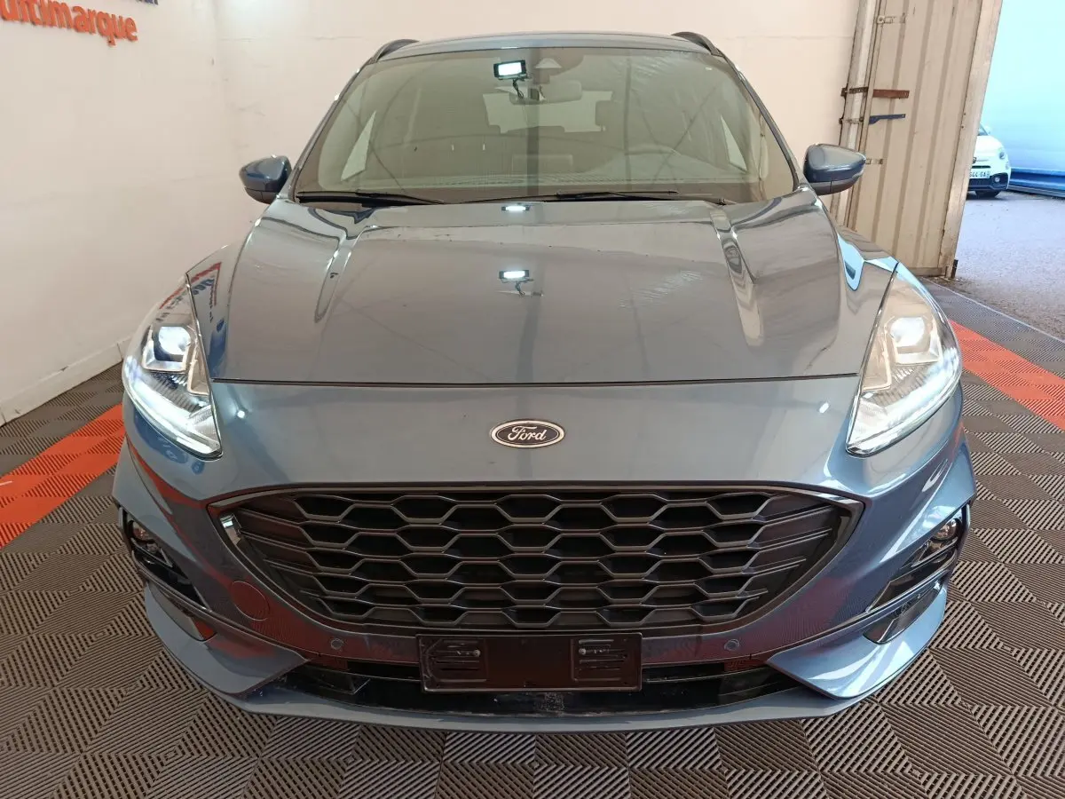 Vue avant rapprochée du Ford Kuga bleu 2024 avec calandre noire et phares allumés dans un intérieur showroom.