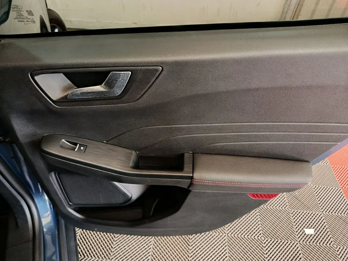 Vue intérieure côté gauche de la porte avant d’un Ford Kuga bleu, avec poignée argentée et accoudoir noir à surpiqûres rouges.