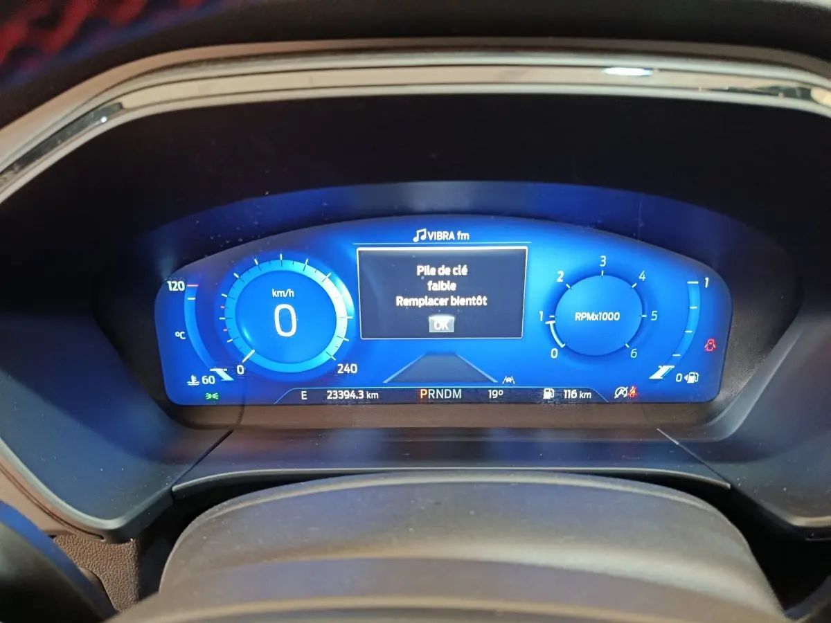 Tableau de bord numérique bleu du Ford Kuga 2024 affichant vitesse, compte-tours et message batterie clé faible.