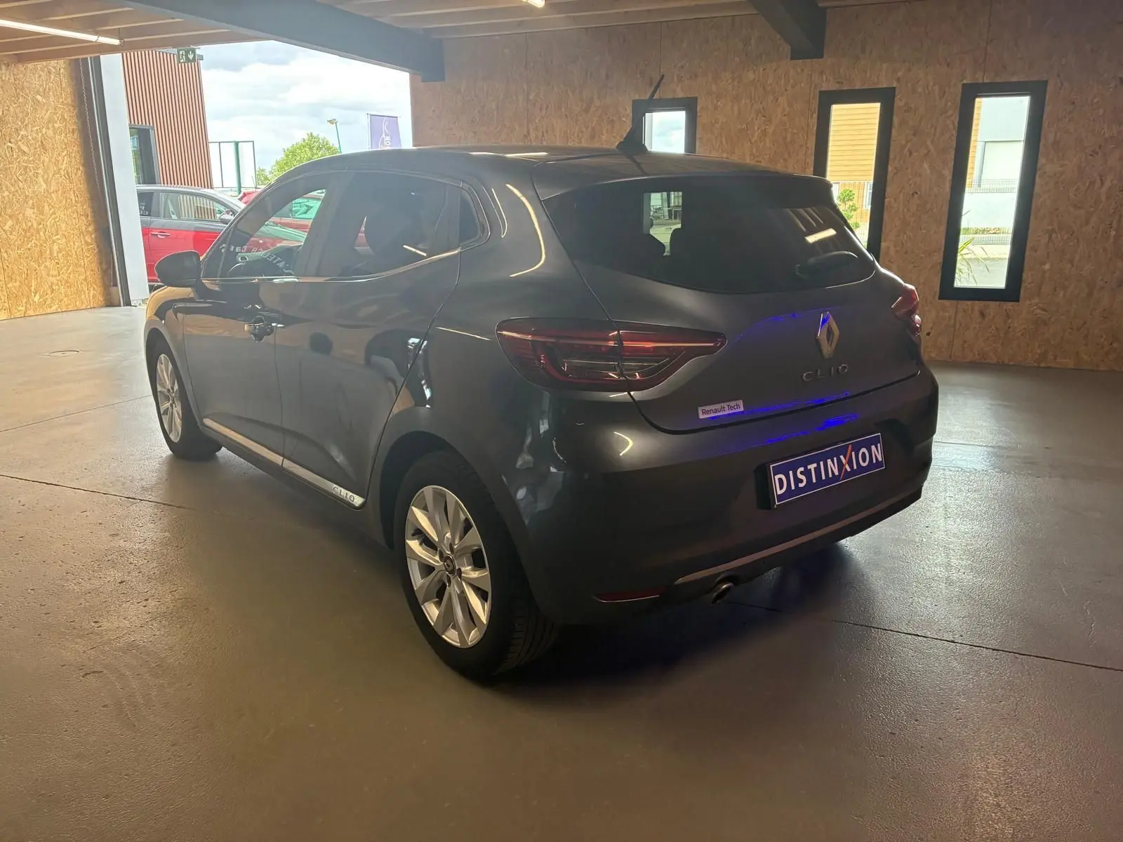 Renault Clio gris 3/4 arrière droit en intérieur, avec logo Renault et plaque Distinxion visible.