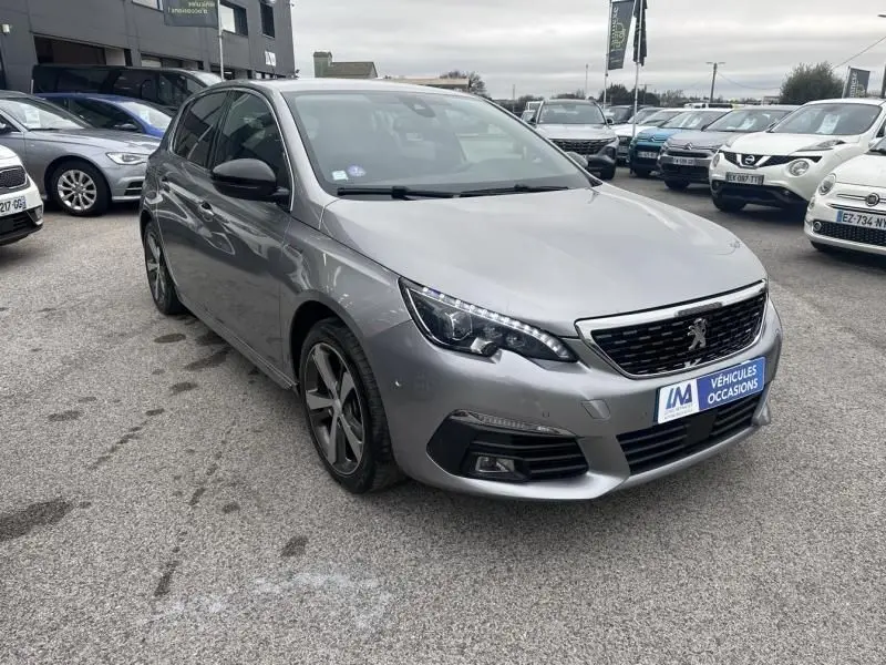 Peugeot 308 gris métal vue 3/4 avant droit, avec calandre noire et jantes alliage distinctives, sur parking extérieur.