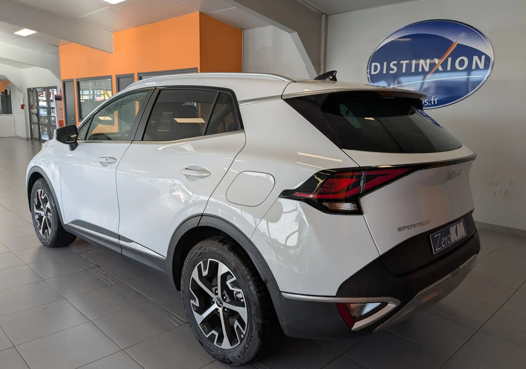 KIA Sportage blanc vue 3/4 arrière droit en intérieur, avec jantes alliage 18’’ et barres de toit visibles.