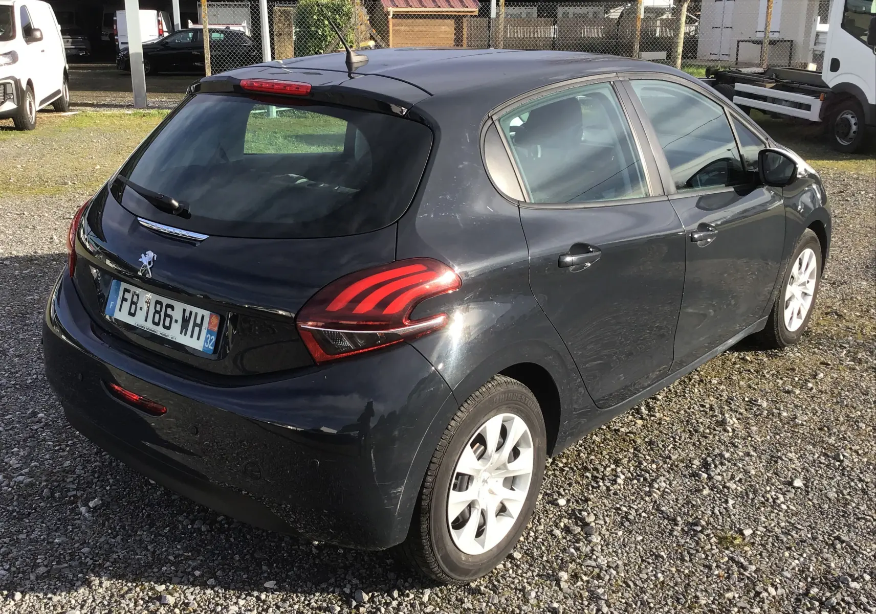 Vue 3/4 arrière droite d'une Peugeot 208 noire 2018 avec feux arrière distinctifs et jantes blanches sur sol gravillonné.
