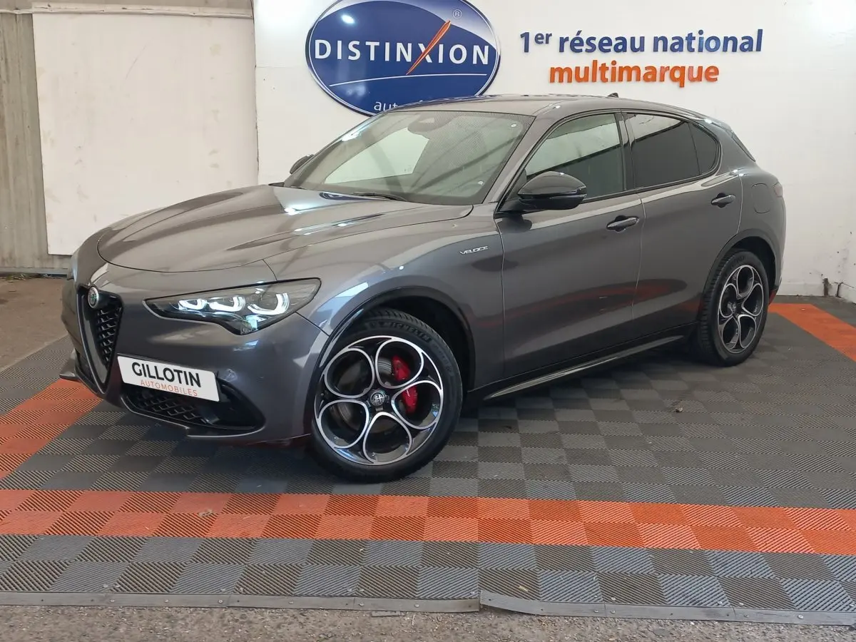 Alfa Romeo Stelvio gris foncé en 3/4 avant droit, avec jantes distinctives et phares LED allumés.