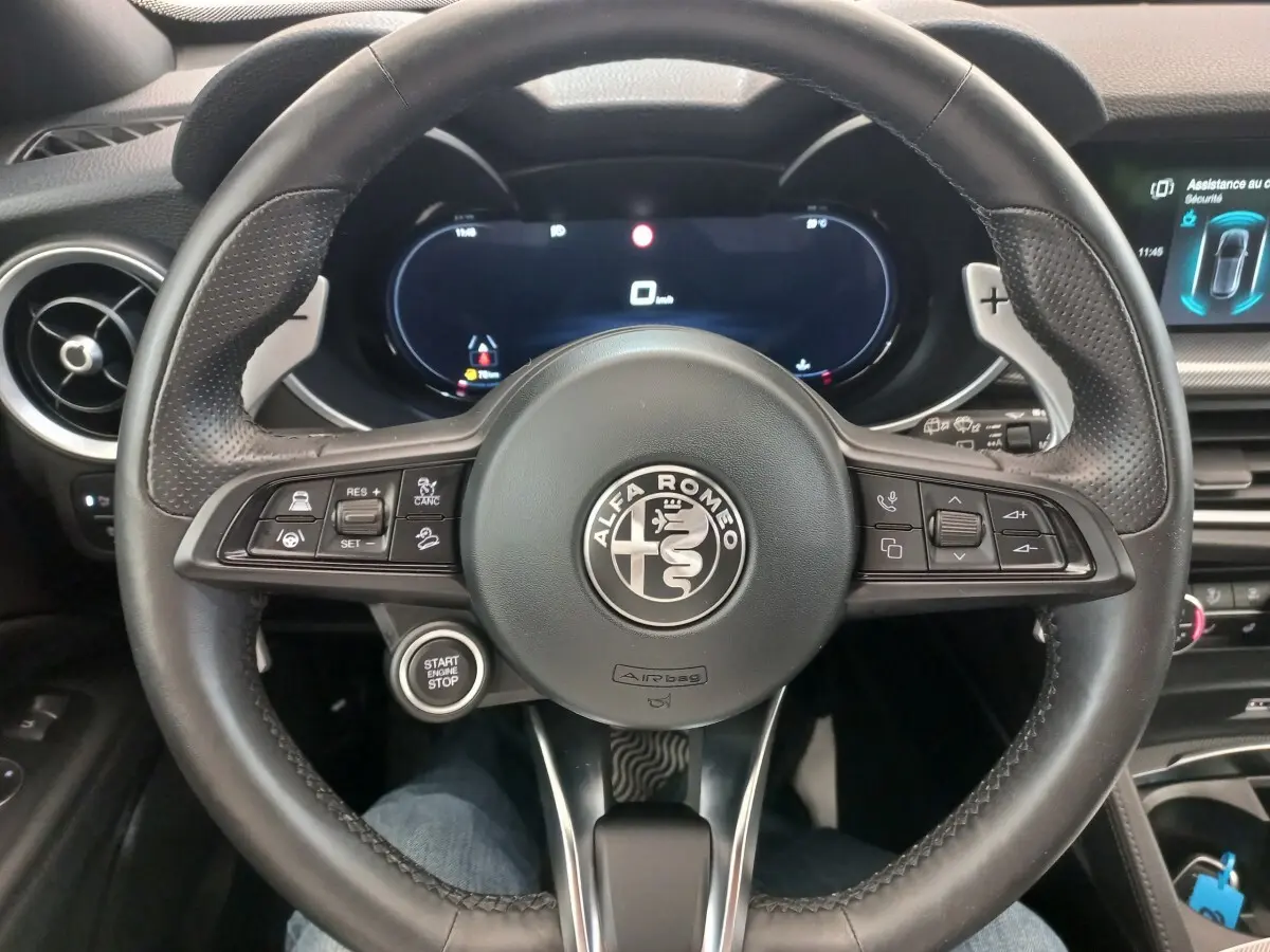Vue rapprochée du volant cuir noir d'une Alfa Romeo Stelvio 2024 avec tableau de bord numérique et bouton start/stop visible.