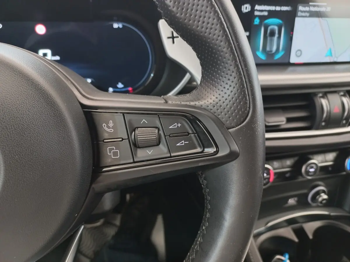 Gros plan sur le volant cuir perforé côté gauche de l'Alfa Romeo Stelvio 2024 avec commandes multifonctions et écran tactile en arrière-plan.