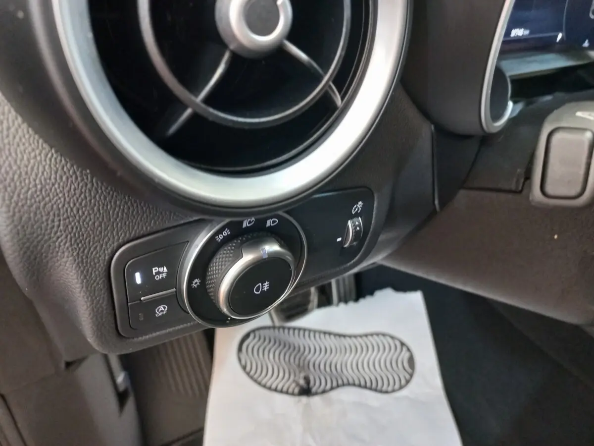 Gros plan sur la commande des phares et l'aération ronde côté gauche du tableau de bord gris foncé de l'Alfa Romeo Stelvio.