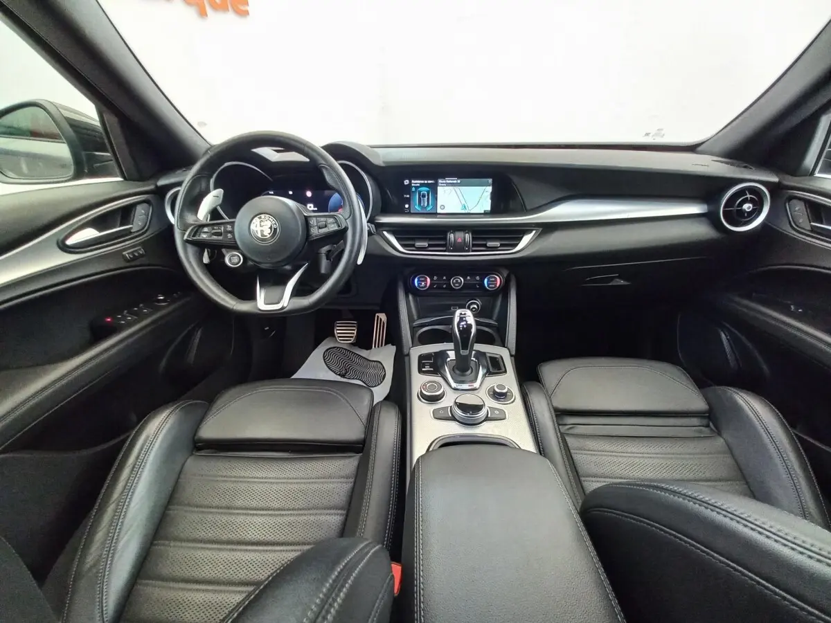 Vue intérieure avant de l'Alfa Romeo Stelvio gris foncé, avec tableau de bord moderne et écran tactile central.