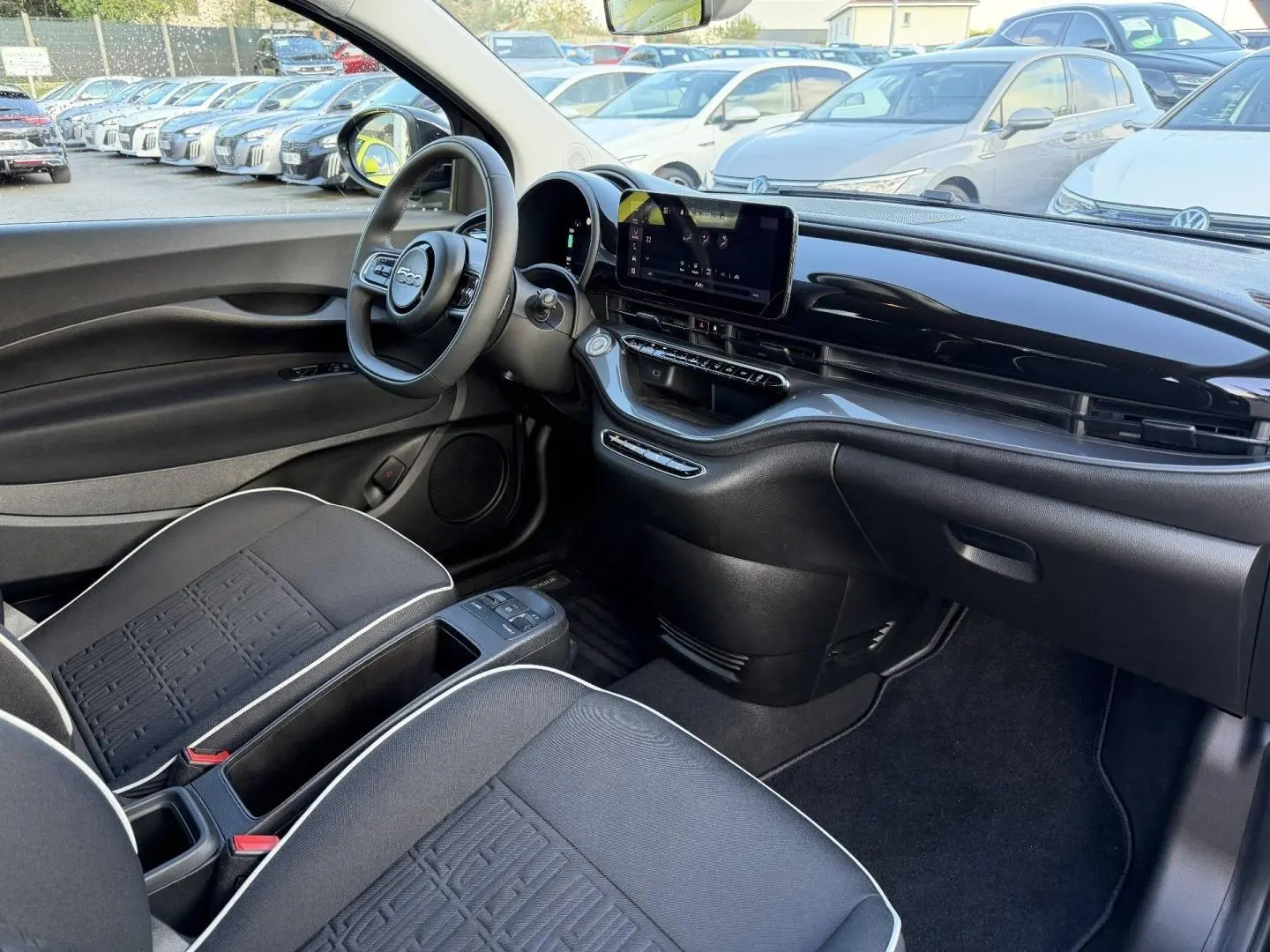 Vue intérieure côté conducteur de la FIAT 500 e 118ch MY24 avec tableau de bord noir brillant et sièges tissu noir monogramme.