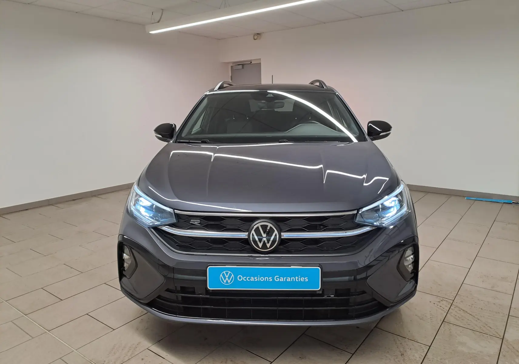 Vue avant d'un Volkswagen Taigo gris cendré avec toit noir et phares LED allumés dans un intérieur neutre.